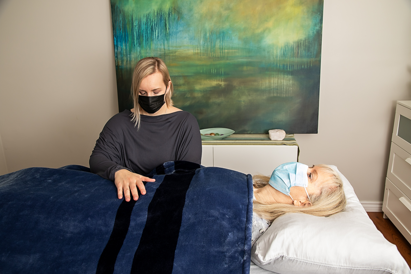 woman receives craniosacral therapy with painting in the background, both wearing masks